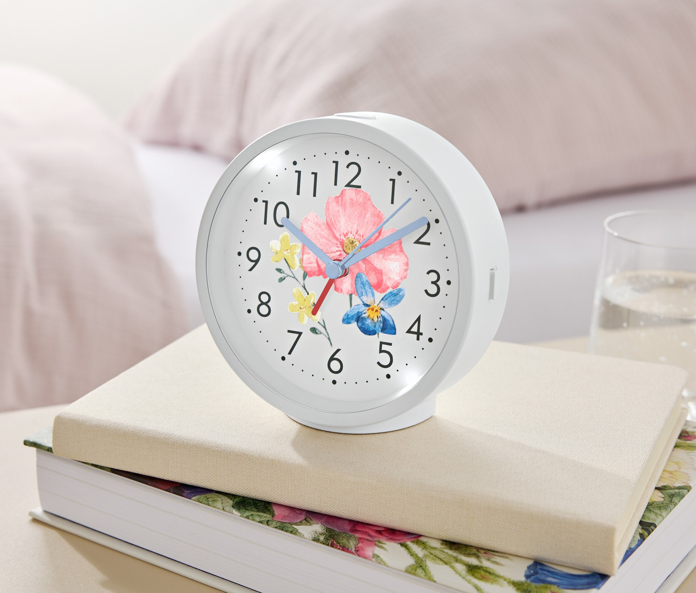 Un réveil blanc avec un motif floral repose sur des livres à côté d'un verre d'eau.