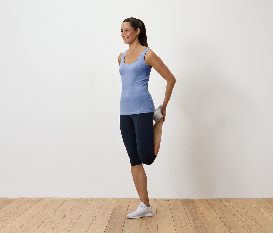 Une femme en débardeur bleu et leggings sombres étire son quadriceps en tenant son pied derrière elle.