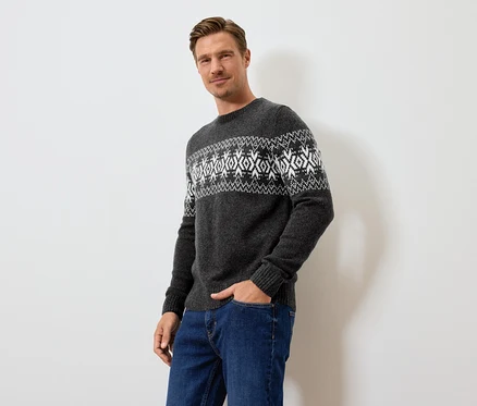 Un homme pose en pull gris à motifs et jean bleu.