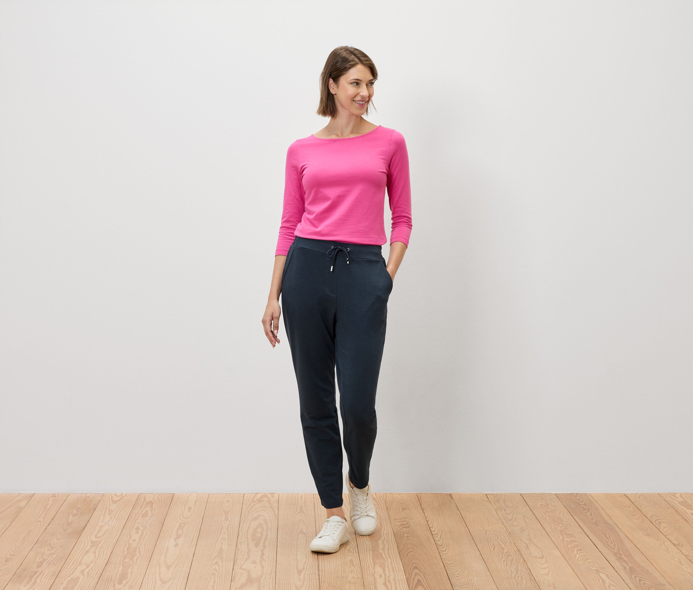 Femme portant un haut rose et un pantalon bleu marine debout sur un plancher en bois devant un mur blanc.