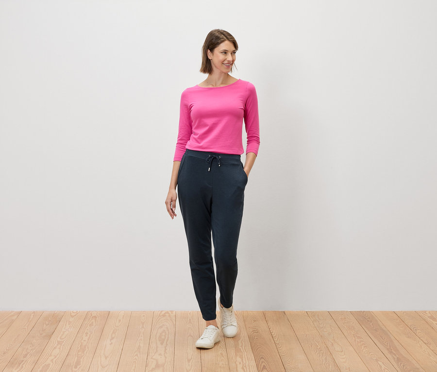 Femme portant un haut rose et un pantalon bleu marine debout sur un plancher en bois devant un mur blanc.