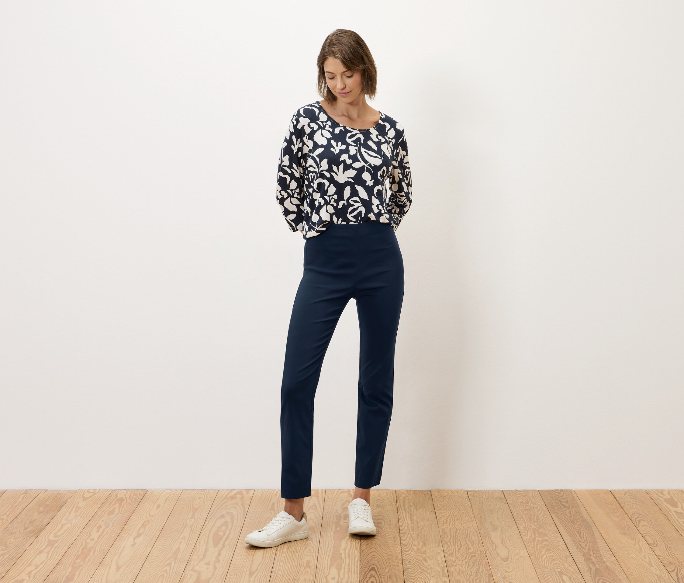 Une femme se tient devant un mur blanc, vêtue d'un pantalon bleu marine et d'un haut à motifs noirs et blancs.