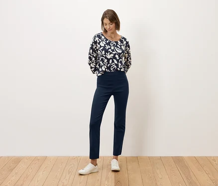Une femme se tient devant un mur blanc, vêtue d'un pantalon bleu marine et d'un haut à motifs noirs et blancs.