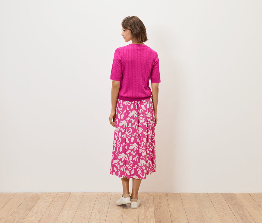Une femme se tient dos à la caméra, elle porte un haut rose et une jupe à motifs floraux.