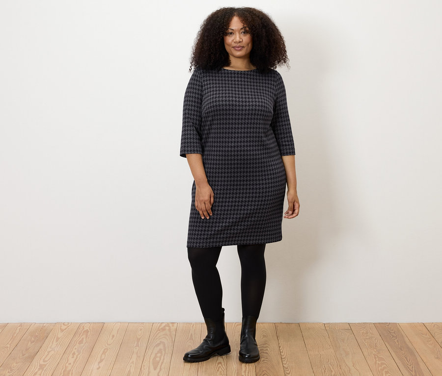 Une femme debout portant une robe en jersey jacquard pied-de-poule, des collants noirs et des bottes noires.