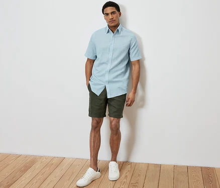 Un homme pose sur la photo et porte une chemise à manches courtes en lin bleu clair, un short kaki et des baskets blanches.