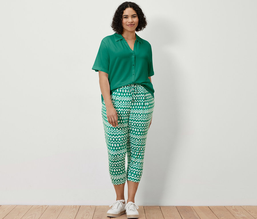 Une femme debout portant un pantalon 3/4 en jersey imprimé et une blouse émeraude avec patte de boutonnage.