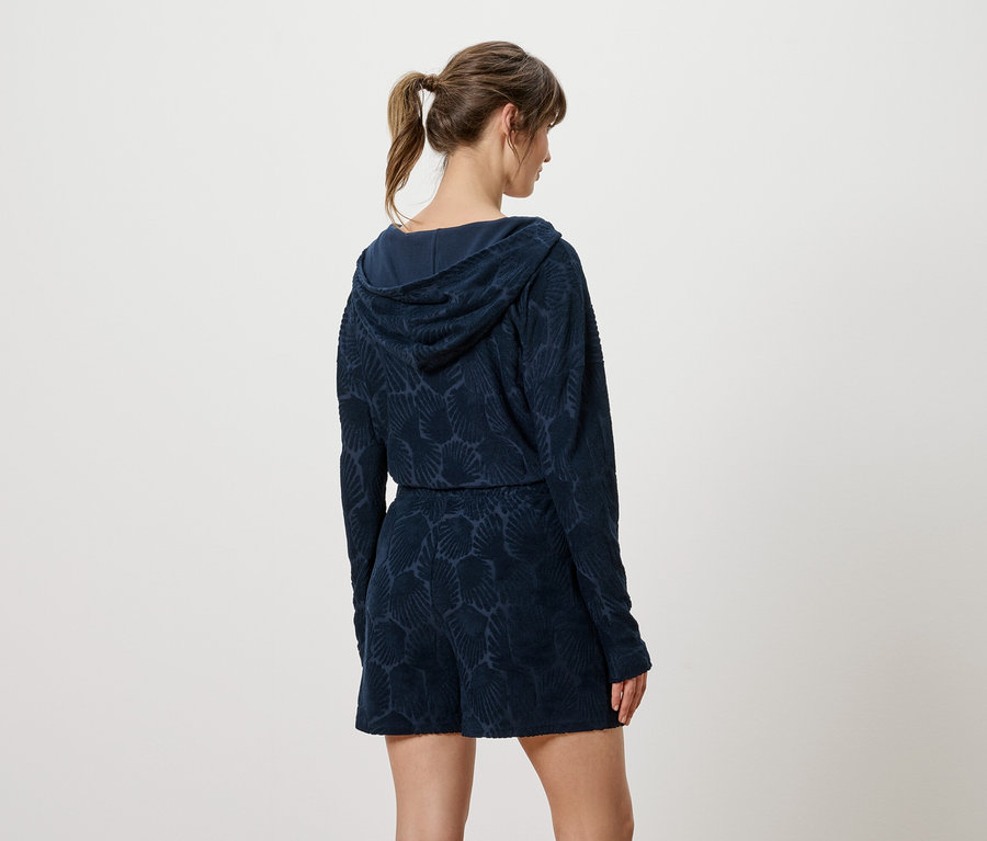 Vue de dos d'une femme aux cheveux bruns attachés en queue de cheval portant un sweat à capuche et un short bleu marine à motifs.