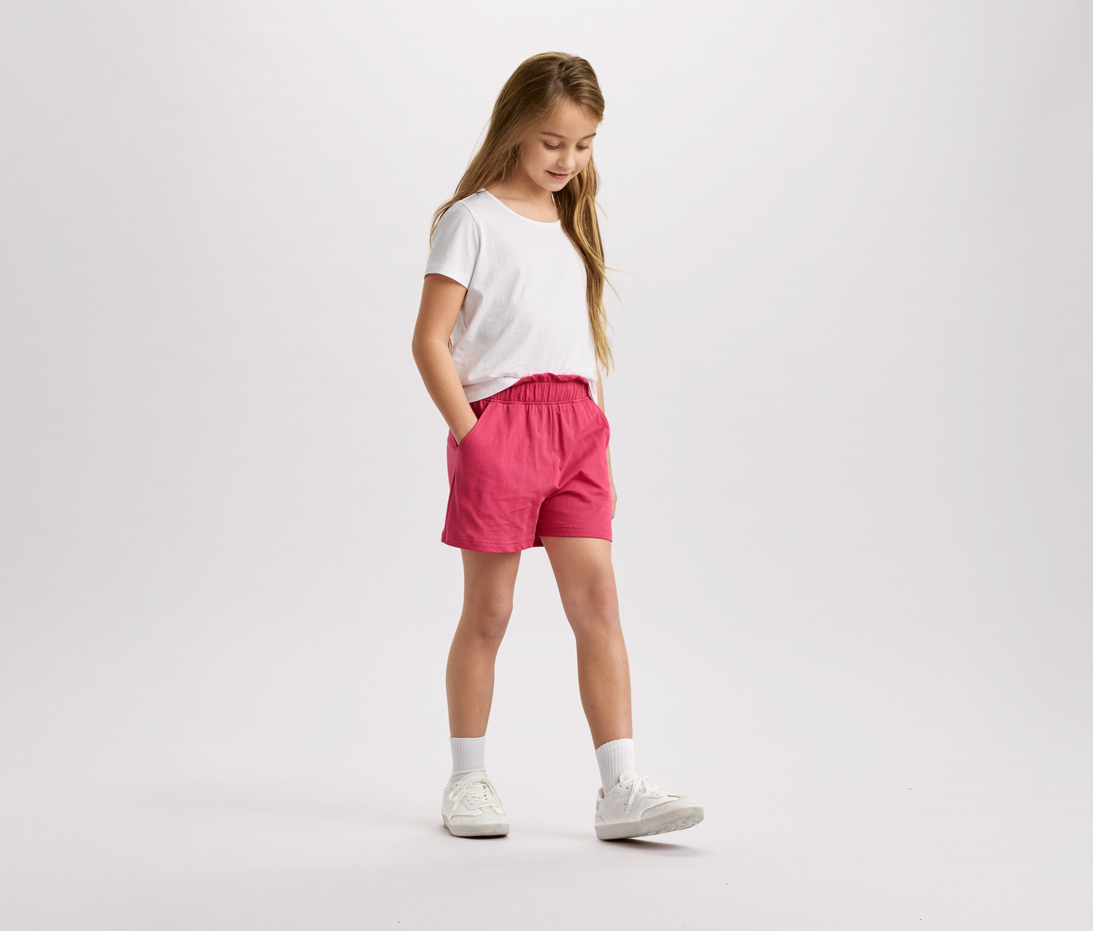 Une fille aux cheveux blonds, portant un t-shirt blanc et un short rose, se tient debout et regarde vers le bas.