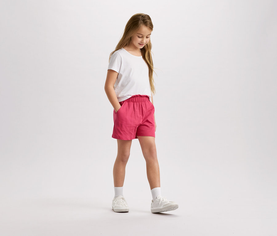 Une fille aux cheveux blonds, portant un t-shirt blanc et un short rose, se tient debout et regarde vers le bas.