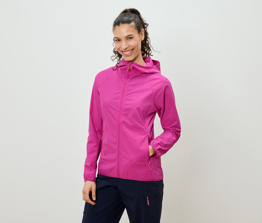 Une femme souriante aux cheveux attachés en queue de cheval porte une veste rose à capuche et un pantalon bleu marine.