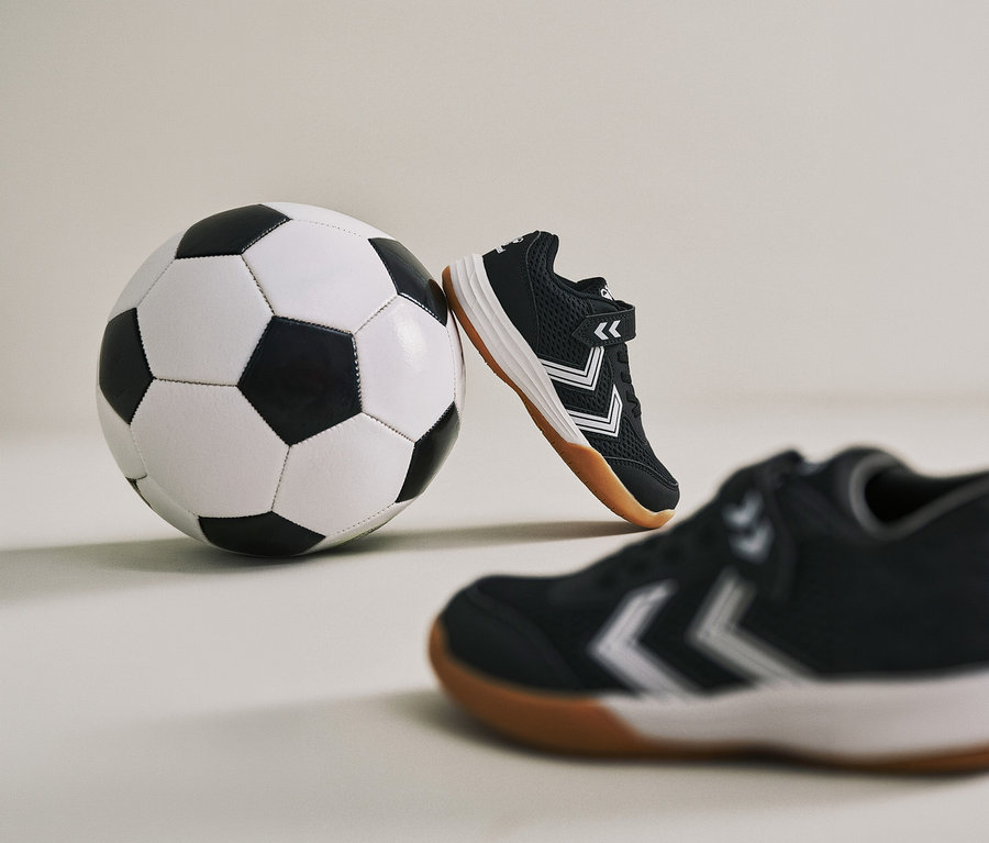 Un ballon de football est appuyé contre une chaussure noire Multiplay Flex VC JR hummel®. Une autre chaussure est visible au premier plan.