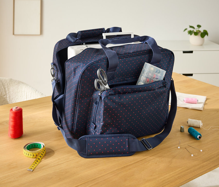Un sac de transport bleu marine à pois rouges est posé sur une table en bois, à côté d'une bobine de fil rouge et d'un ruban à mesurer jaune.