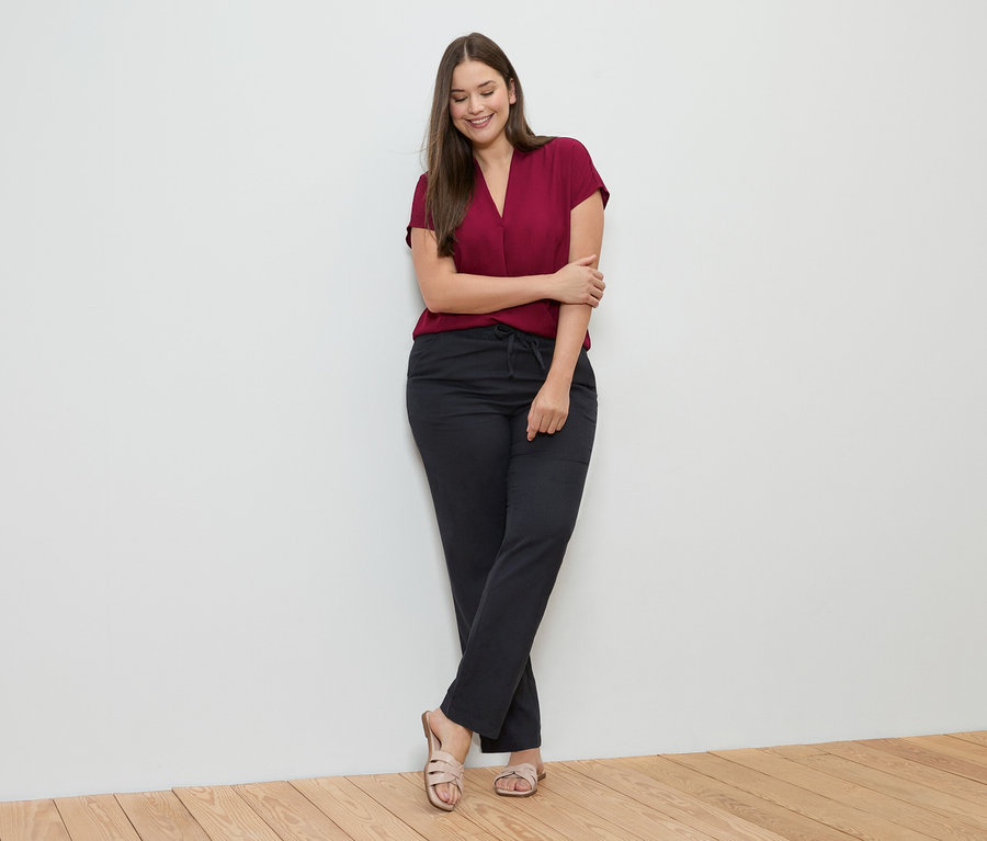 Femme appuyée contre un mur, portant un haut bordeaux, un pantalon en lin noir et des sandales.