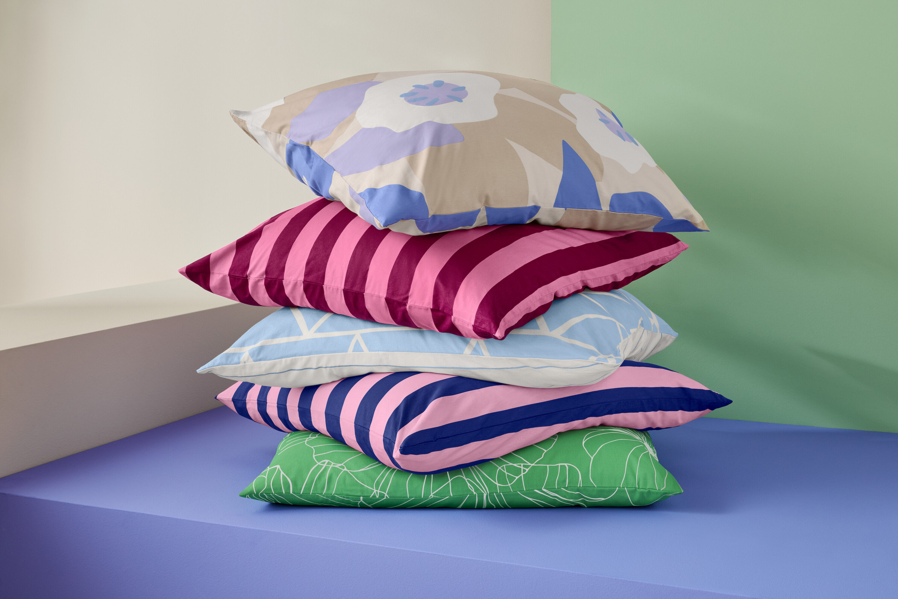Une pile de cinq coussins sur un fond violet devant un fond beige et vert.