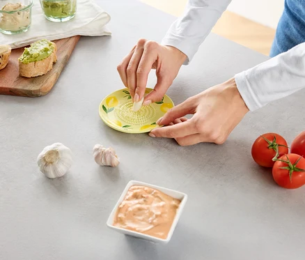 Une personne râpe de l'ail sur une râpe jaune, entourée de tomates et de sauces.