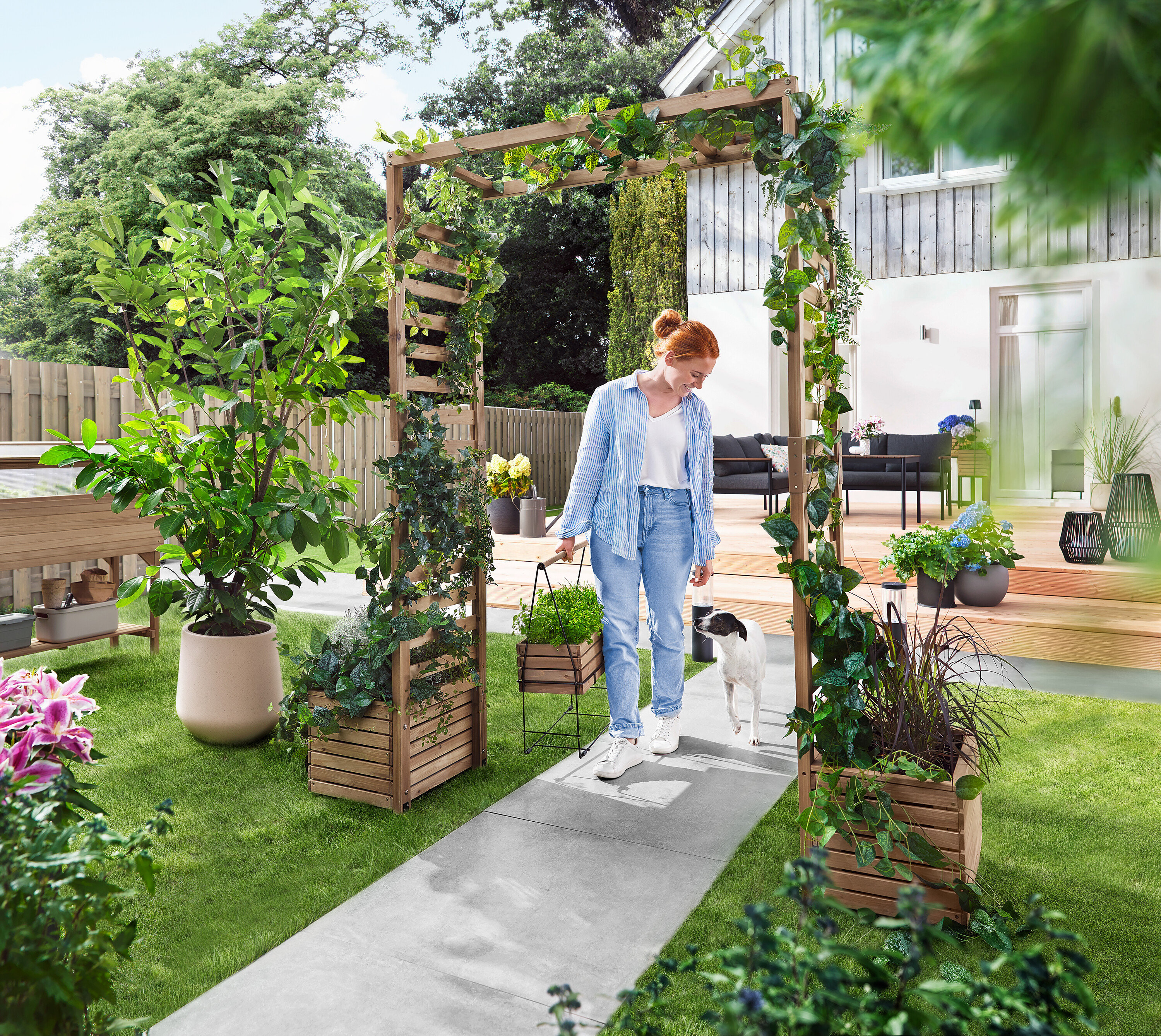 Une femme marche avec un chien à travers une arche de jardin recouverte de plantes. Elle porte un bac à plantes.
