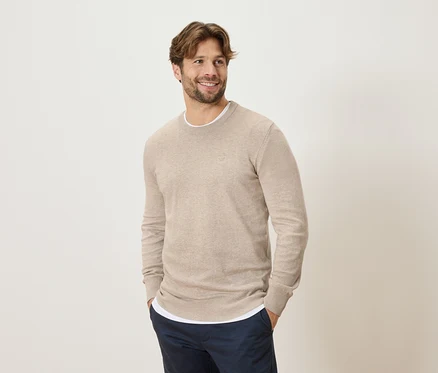 Homme souriant portant un pull beige et un pantalon bleu, les mains dans les poches, regardant sur le côté.