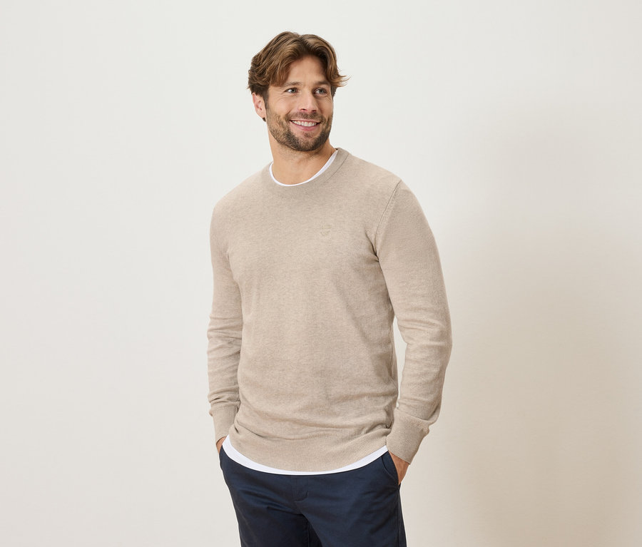 Homme souriant portant un pull beige et un pantalon bleu, les mains dans les poches, regardant sur le côté.