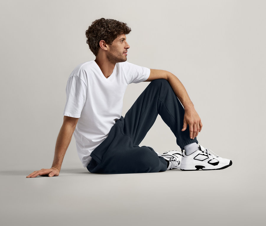 Un homme aux cheveux bouclés est assis par terre, portant un t-shirt blanc, un pantalon de survêtement bleu marine et des baskets blanches et noires.