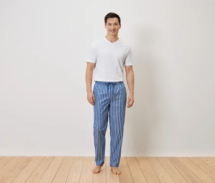 Un homme se tient pieds nus en pantalon de pyjama rayé bleu et blanc et en t-shirt blanc devant un mur blanc.