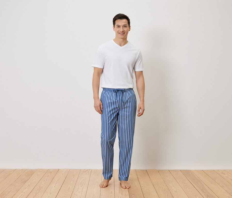 Un homme se tient pieds nus en pantalon de pyjama rayé bleu et blanc et en t-shirt blanc devant un mur blanc.