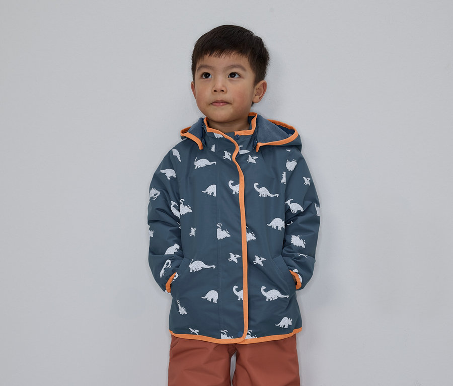 Un garçon aux cheveux foncés porte une veste bleue à motifs de dinosaures avec des détails orange sur un fond gris.