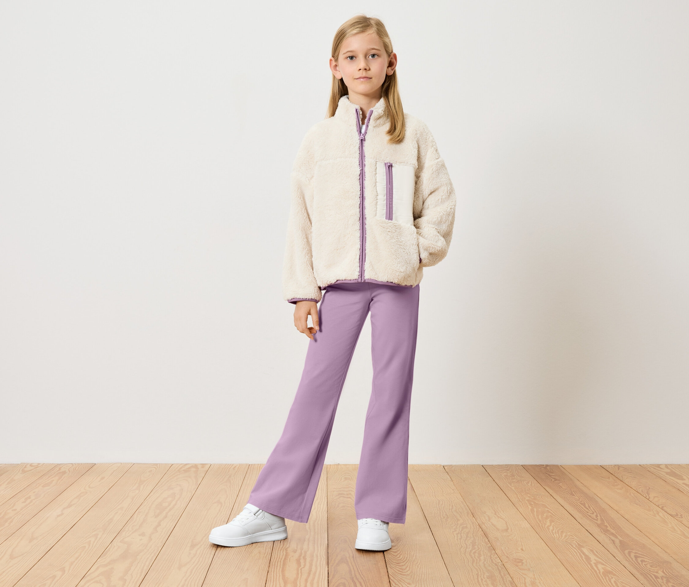 Fillette portant une veste teddy beige avec des détails violets, un pantalon violet et des baskets blanches, debout devant un mur blanc.