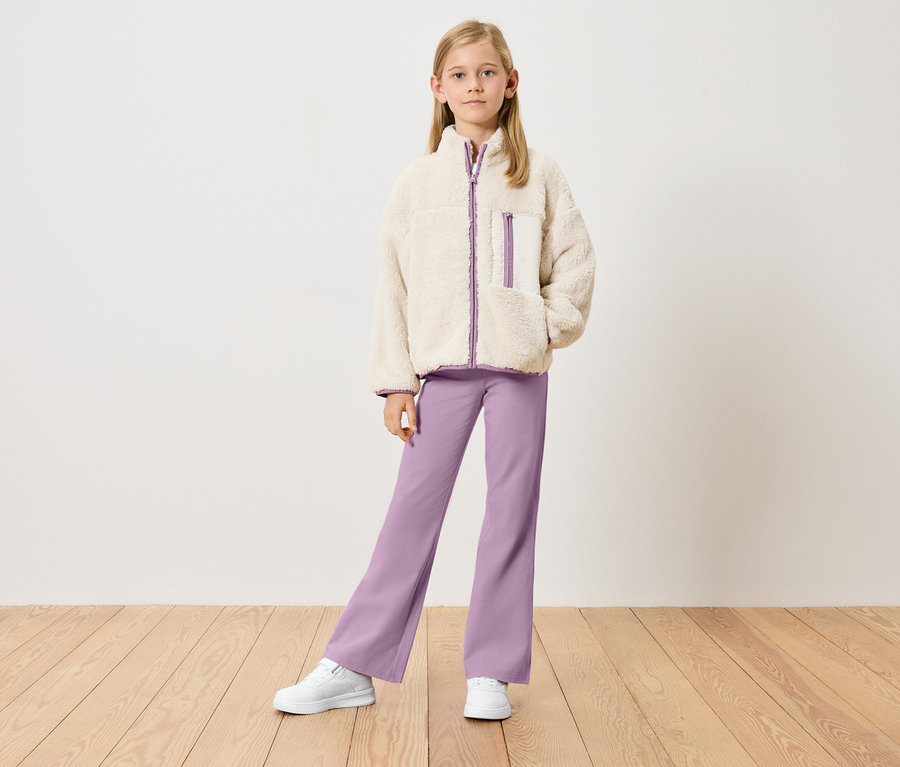 Fillette portant une veste teddy beige avec des détails violets, un pantalon violet et des baskets blanches, debout devant un mur blanc.