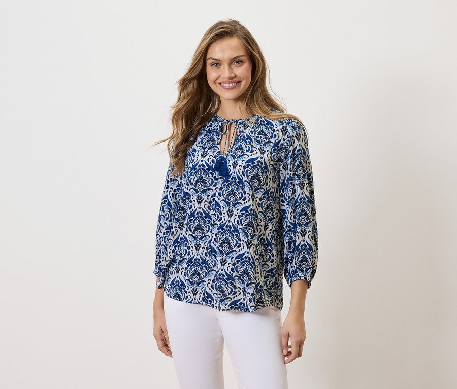 Une femme aux longs cheveux blonds, souriant, portant un chemisier à motifs bleus et blancs et un pantalon blanc.