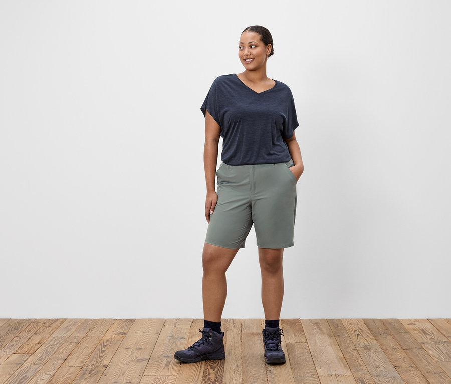 Une femme se tient debout, les mains dans les poches, portant un t-shirt bleu marine, un short vert et des chaussures de randonnée.