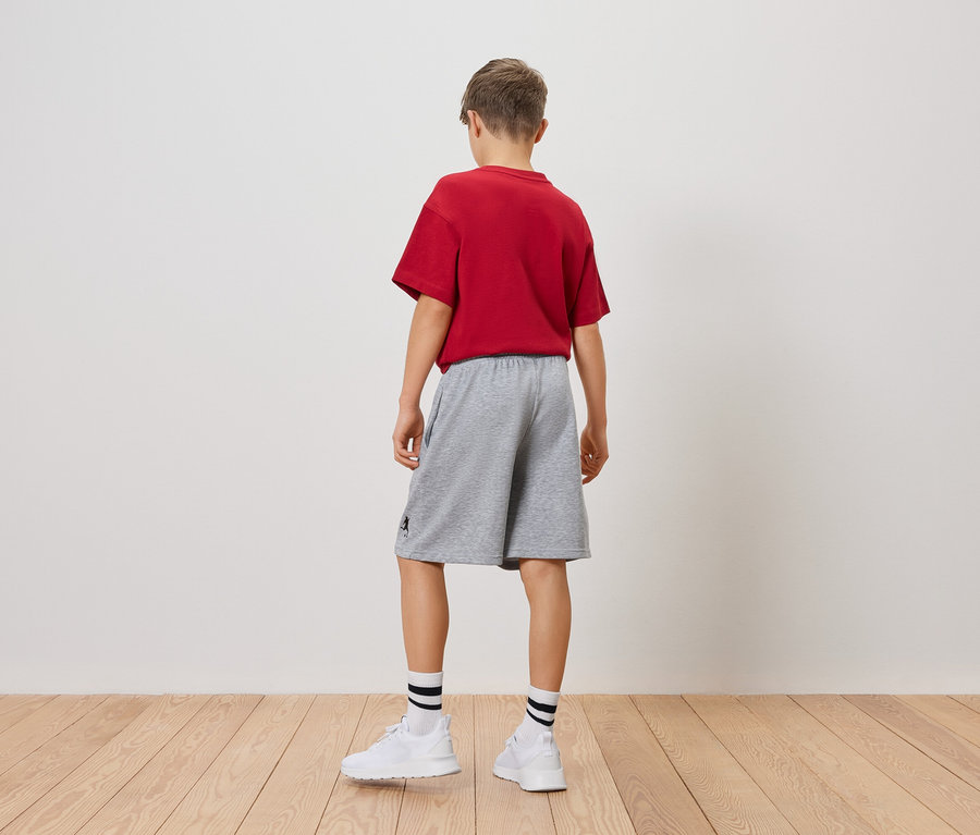 Un jeune garçon se tient dos à la caméra, il porte un t-shirt rouge, un short gris, des chaussettes blanches et des baskets.