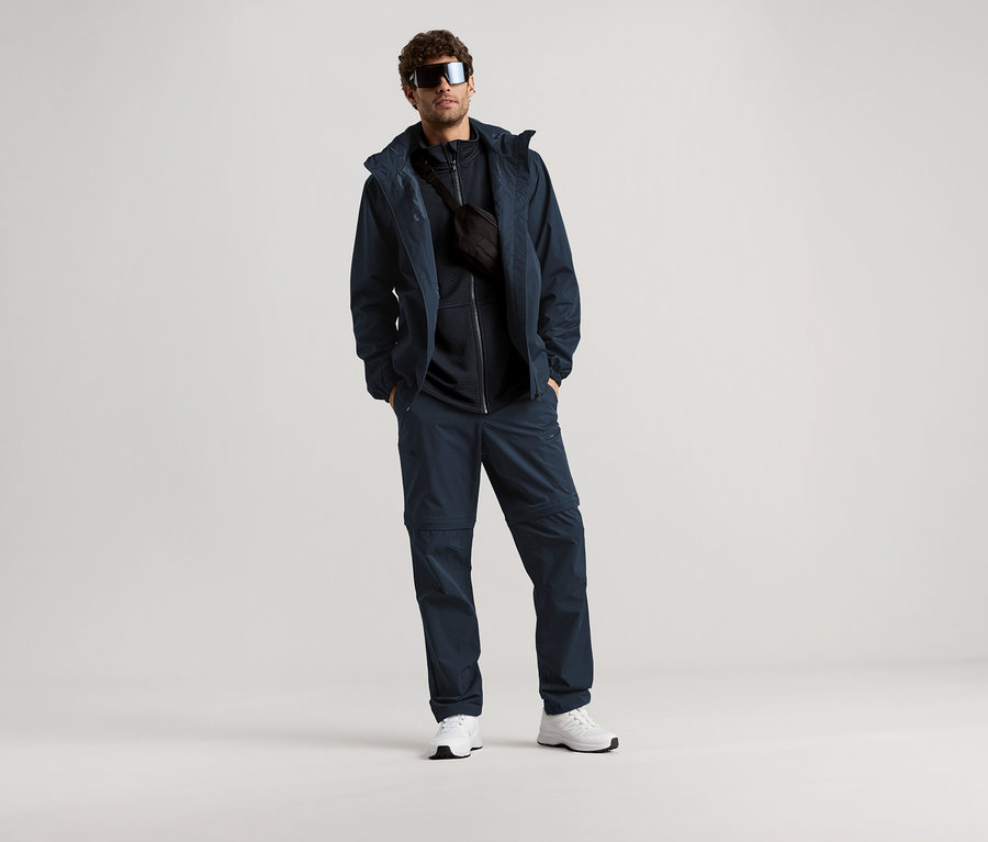 Un homme se tient debout avec des lunettes de soleil, une veste bleu marine, un pantalon et un sac banane sur un fond clair.