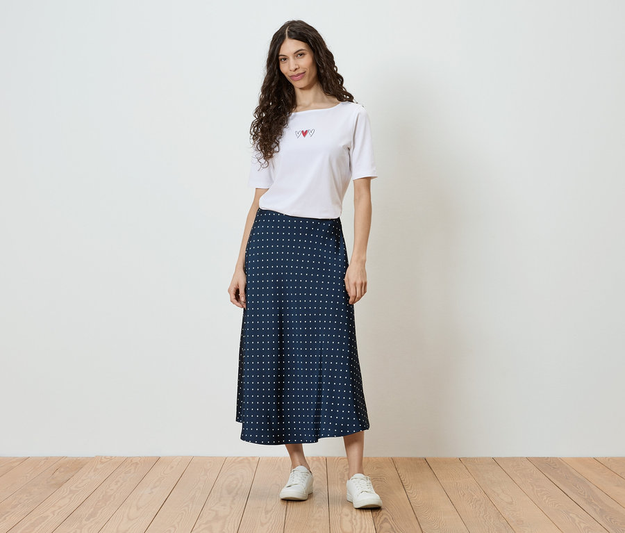 Une femme aux cheveux bouclés porte un t-shirt blanc avec un motif de cœur et une jupe bleu marine à pois blancs, ainsi que des baskets blanches.