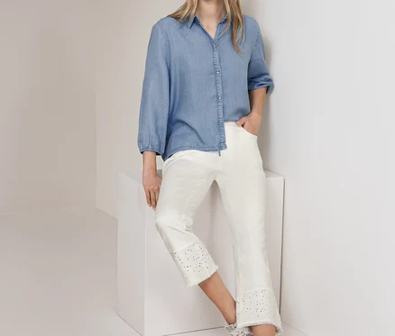Femme debout, portant un chemisier en jean Street One bleu clair et un jean Street One blanc court avec broderie anglaise, appuyée contre un mur et un cube blancs.