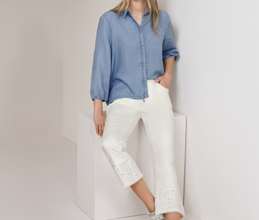 Femme debout, portant un chemisier en jean Street One bleu clair et un jean Street One blanc court avec broderie anglaise, appuyée contre un mur et un cube blancs.