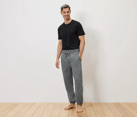 Un homme en t-shirt noir et pantalon de jogging gris, pieds nus sur un plancher en bois devant un mur blanc.