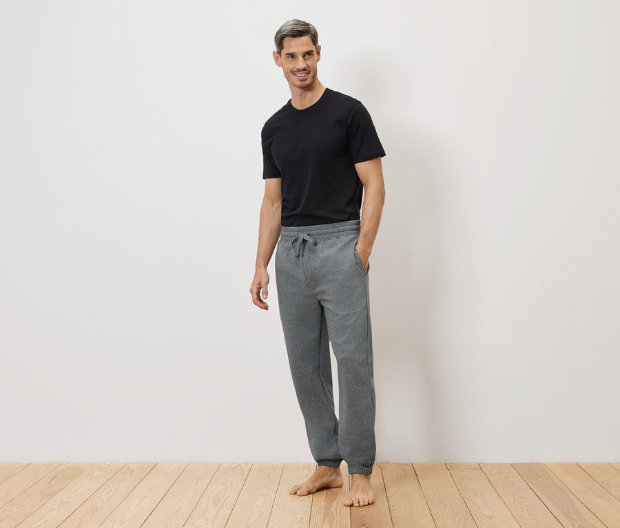 Un homme en t-shirt noir et pantalon de jogging gris, pieds nus sur un plancher en bois devant un mur blanc.