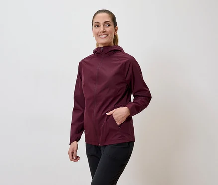 Une femme souriante portant une veste bordeaux à capuche et un pantalon noir, la main dans la poche.
