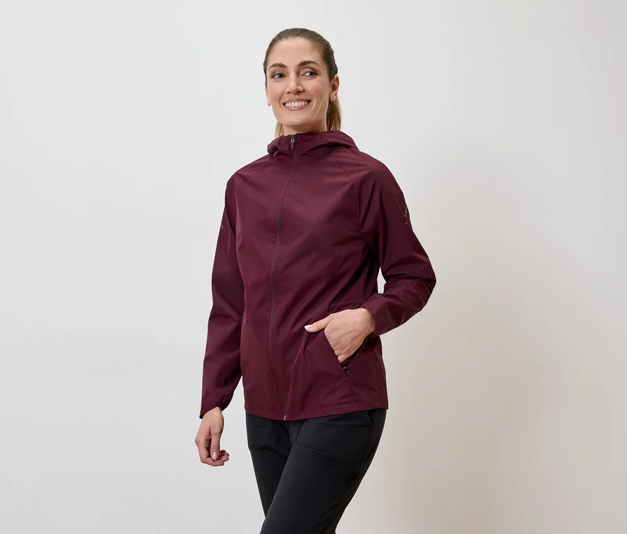 Une femme souriante portant une veste bordeaux à capuche et un pantalon noir, la main dans la poche.