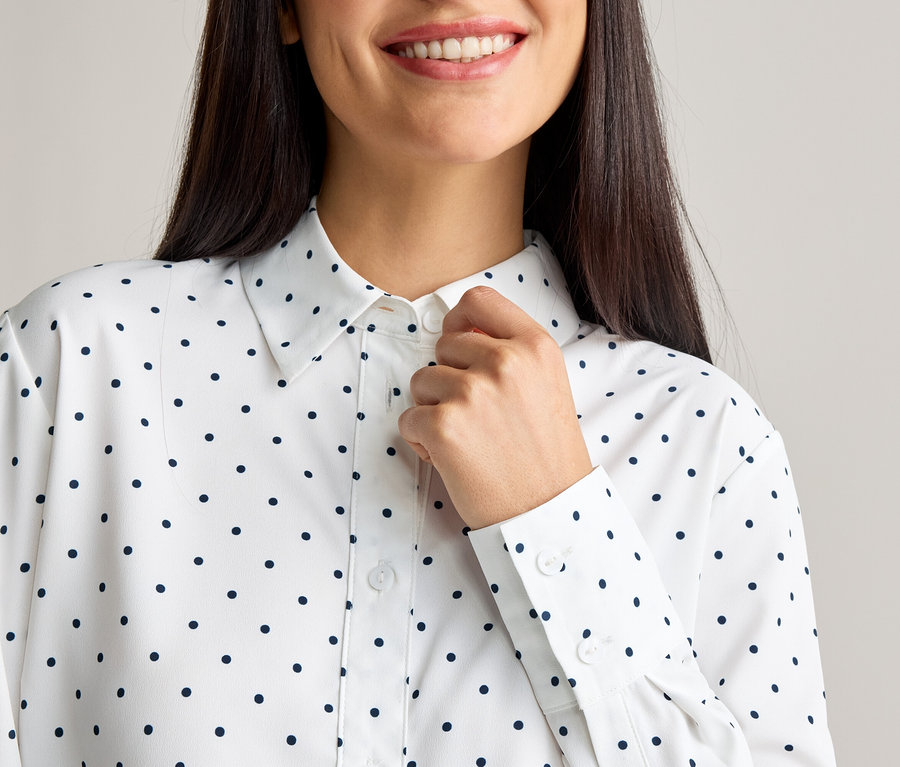 Une femme boutonne le bouton supérieur d'un chemisier blanc à pois noirs.