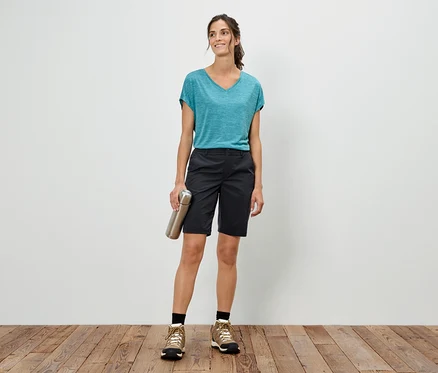 Femme debout devant un mur blanc, tenant une bouteille argentée, portant un t-shirt bleu, un short et des chaussures de randonnée.