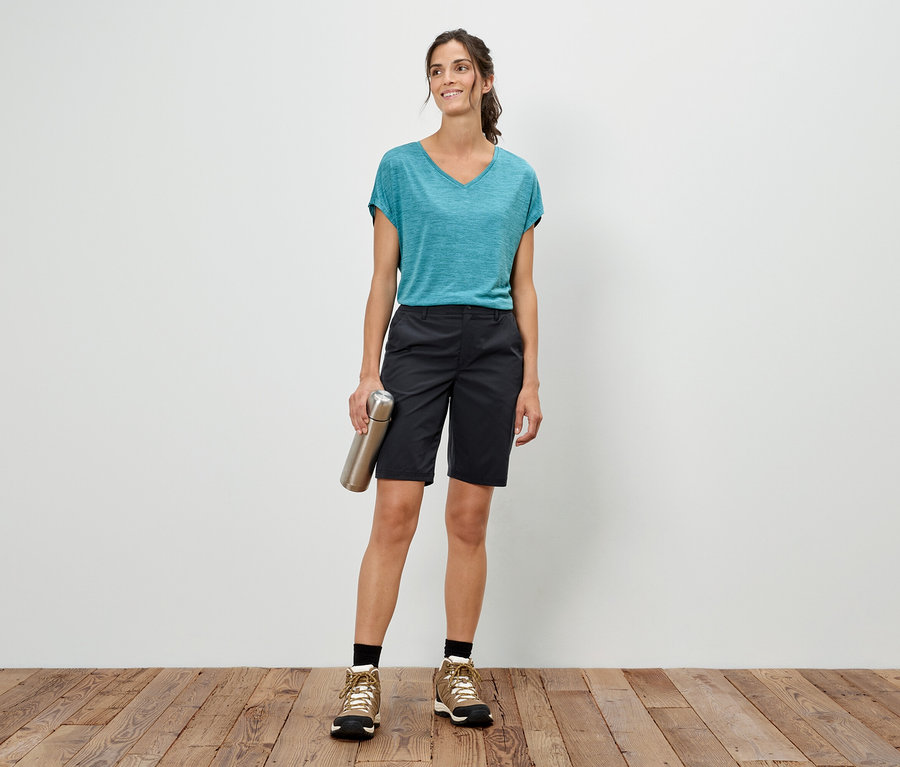 Femme debout devant un mur blanc, tenant une bouteille argentée, portant un t-shirt bleu, un short et des chaussures de randonnée.