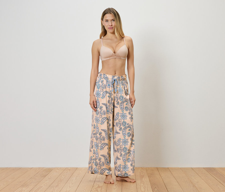 Une femme se tient pieds nus en soutien-gorge et un pantalon ample rose clair avec un motif floral bleu devant un mur.