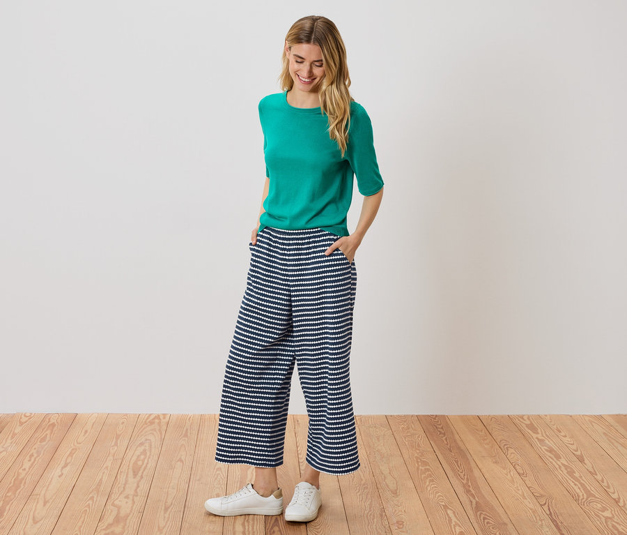 Une femme se tient debout, les mains dans les poches d'une culotte rayée bleu et blanc, portant un haut vert et des baskets blanches.