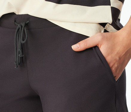 Gros plan d'une personne portant un short de pyjama SCHIESSER gris foncé avec un cordon de serrage noué et une main dans la poche droite. Un haut rayé noir et écru est visible au-dessus du short.