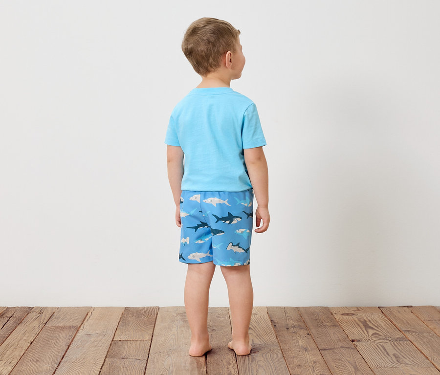 Vue de dos d'un garçon portant un t-shirt bleu clair et un short de bain avec un motif de requins, debout sur un plancher en bois devant un mur blanc.