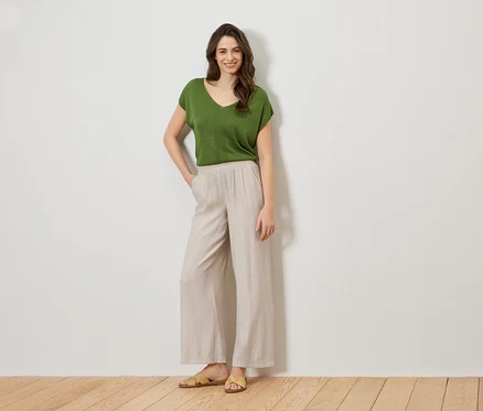 Femme debout, la main dans la poche, portant un haut vert et un pantalon beige.