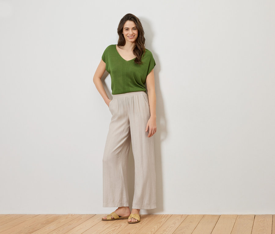 Femme debout, la main dans la poche, portant un haut vert et un pantalon beige.