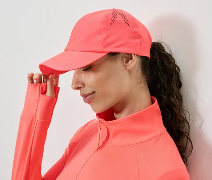 Une femme portant une veste et une casquette corail touche le bord de la casquette.